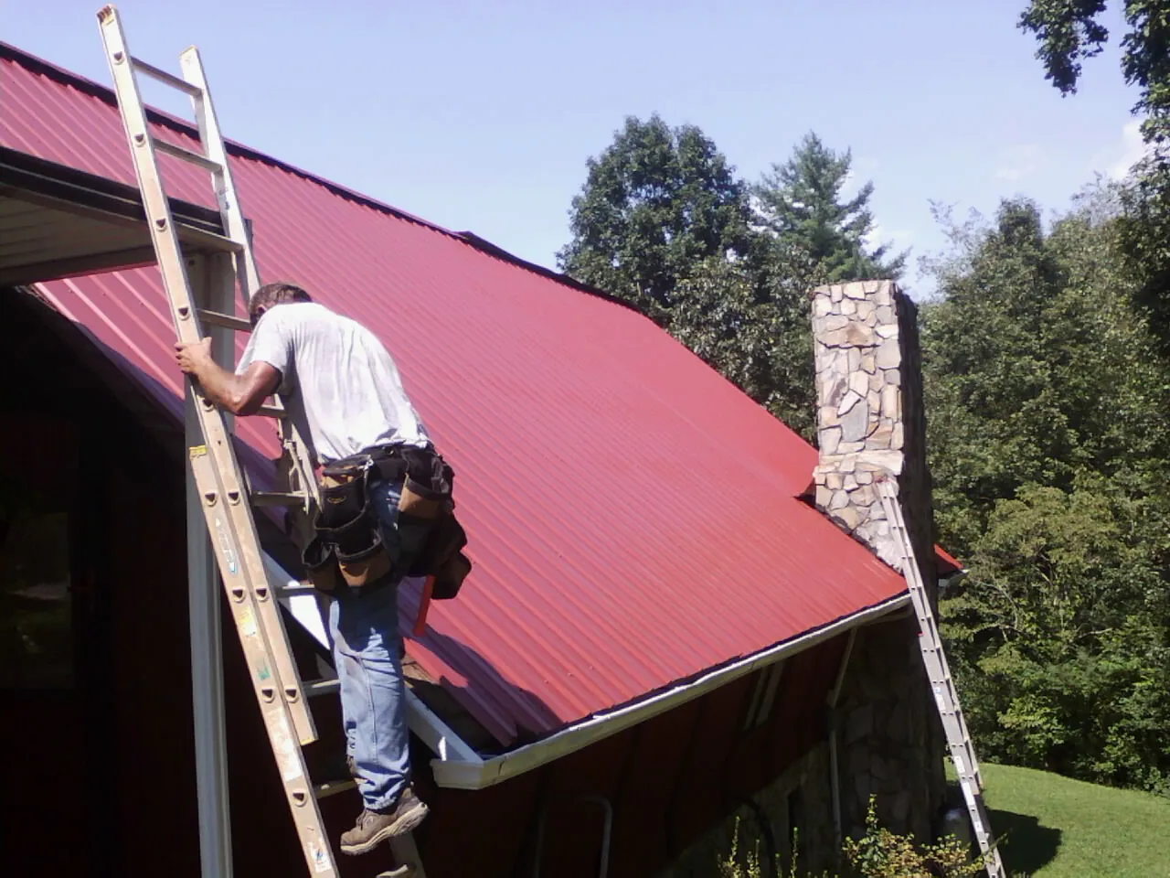 Roofer on a ladder beside a red metal roof and stone chimney — Soffit & Fascia Repair by Roofing Brothers in Boone